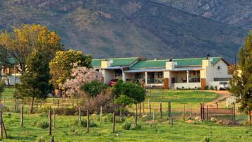 Una casa con techo verde, rodeada por un campo cercado, y una montaña al fondo.