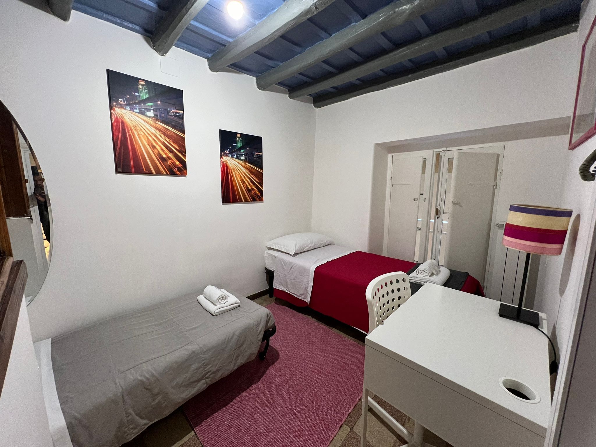 A small, single-room hotel room with a bed, a desk, and two night-time cityscape photographs on the wall.