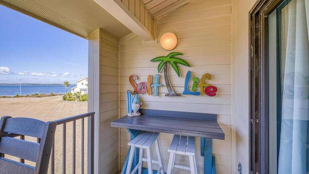 Un balcon donnant sur la plage, un mur en bois orné d’une enseigne « Salt Life », et une petite table avec des tabourets.