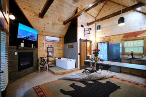 A cozy cabin interior with a fireplace, a large TV, a bathtub, and a kitchen area.