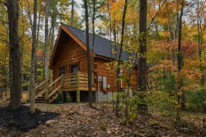 A log cabin with a wooden staircase leading to a deck, surrounded by trees with autumn foliage.