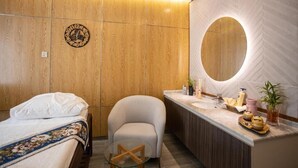 Une chambre d’hôtel avec un mur lambrissé en bois, un miroir rond, un lit recouvert d’une courtepointe à motifs floraux, un petit fauteuil et une coiffeuse avec des articles de toilette.