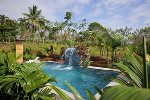 A swimming pool with a waterfall feature surrounded by lush greenery and palm trees. - Root Wellness Arenal (La Tigra)
