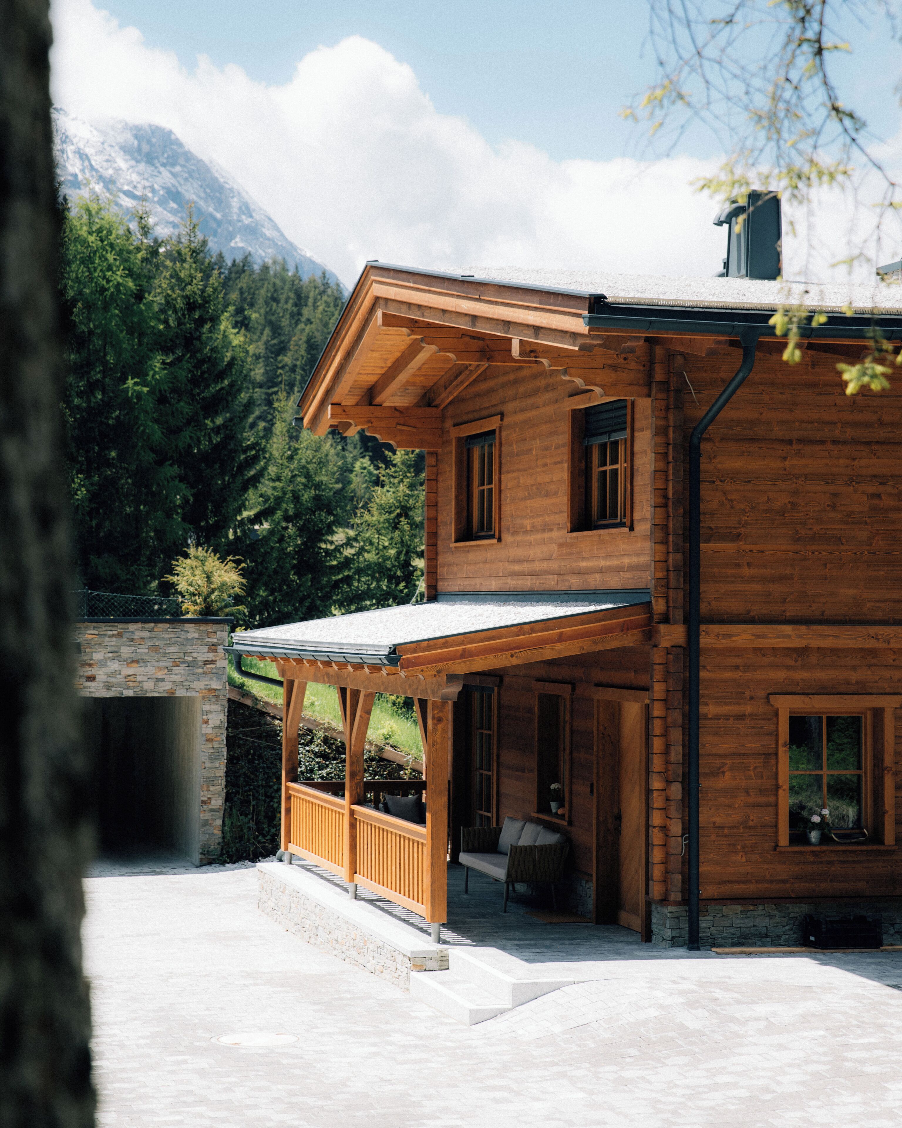 A two-story wooden house with a stone foundation, a covered porch, and a mountain backdrop.