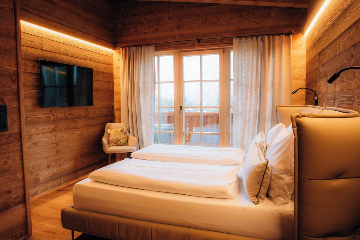 A wooden cabin-style room with a bed, a chair, a TV mounted on the wall, and a window with curtains.