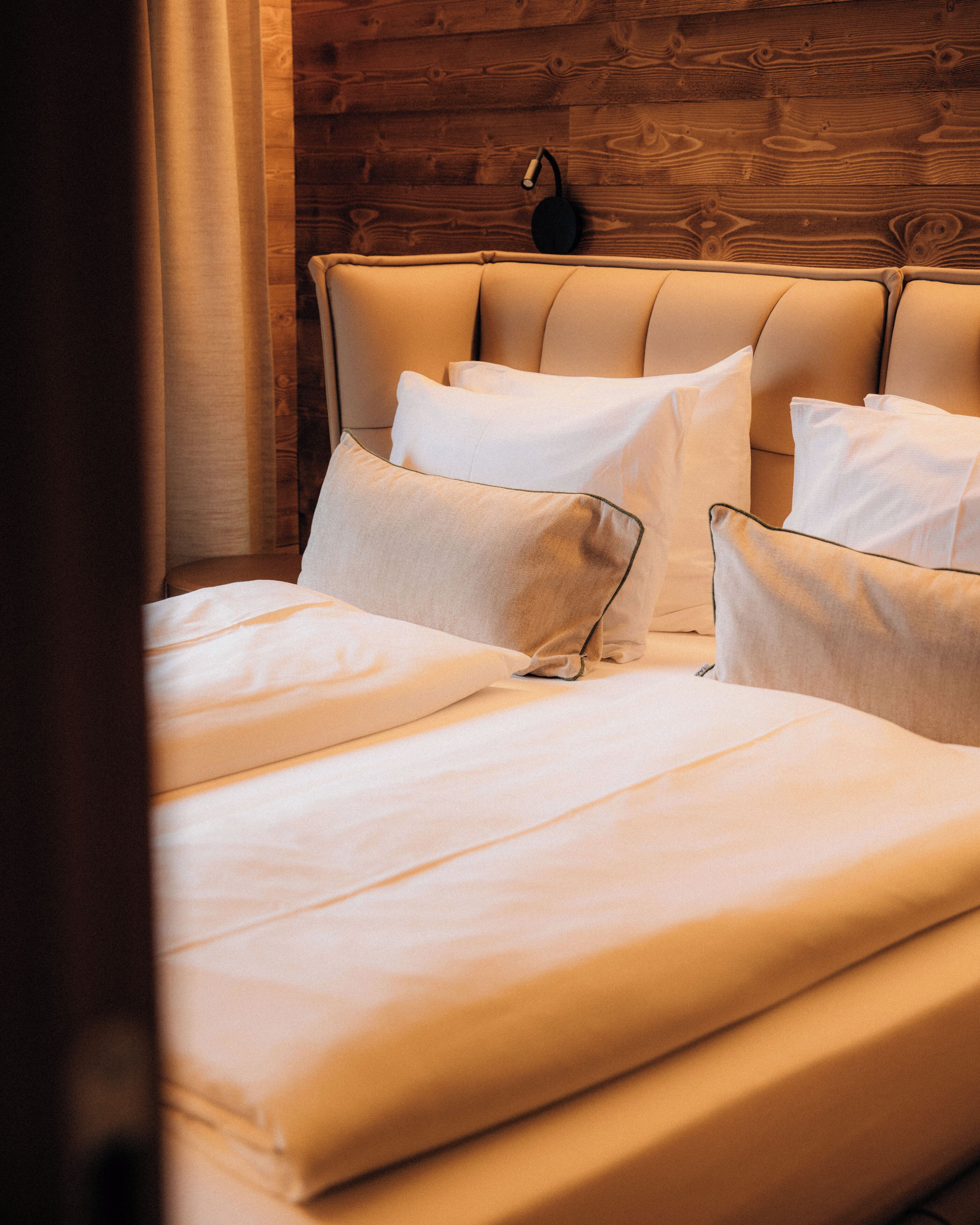 A neatly made bed with white linens and pillows, a wooden headboard, and a wall-mounted light fixture.