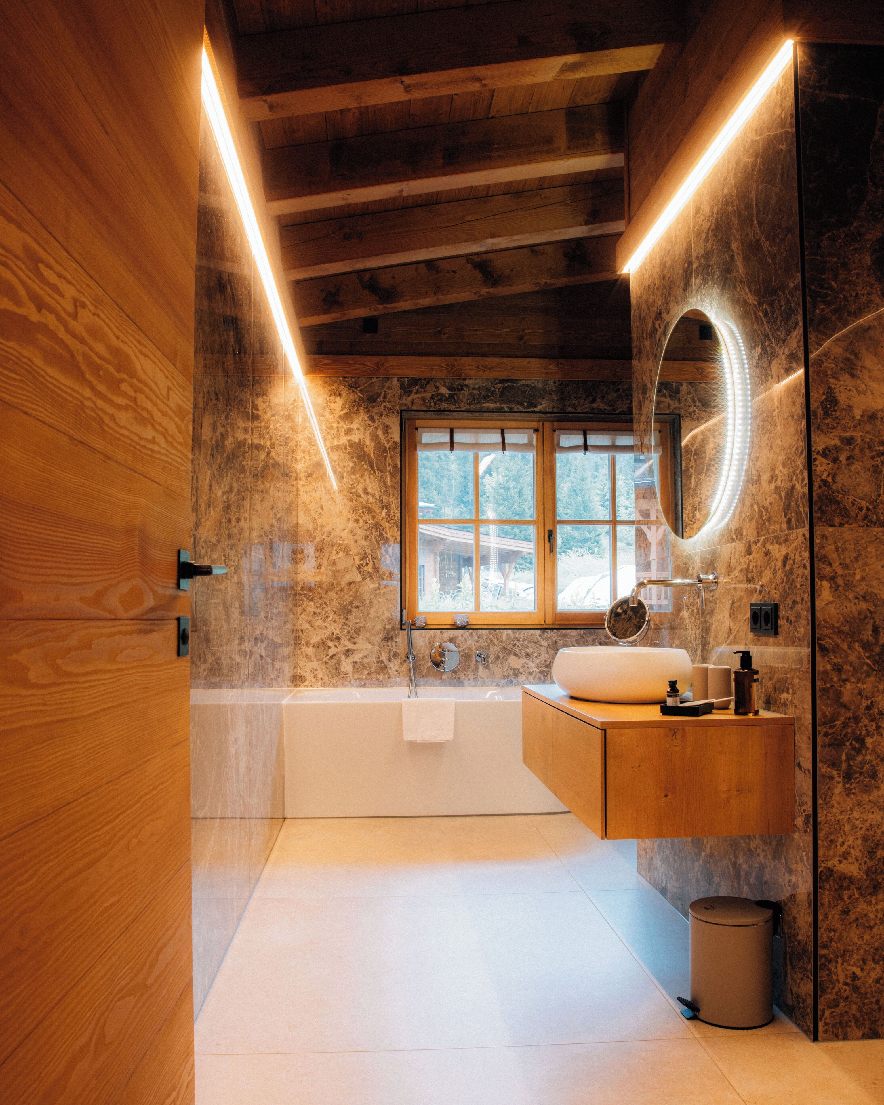 Un baño moderno con tocador de madera, una bañera independiente, un espejo grande con luz y una ventana.
