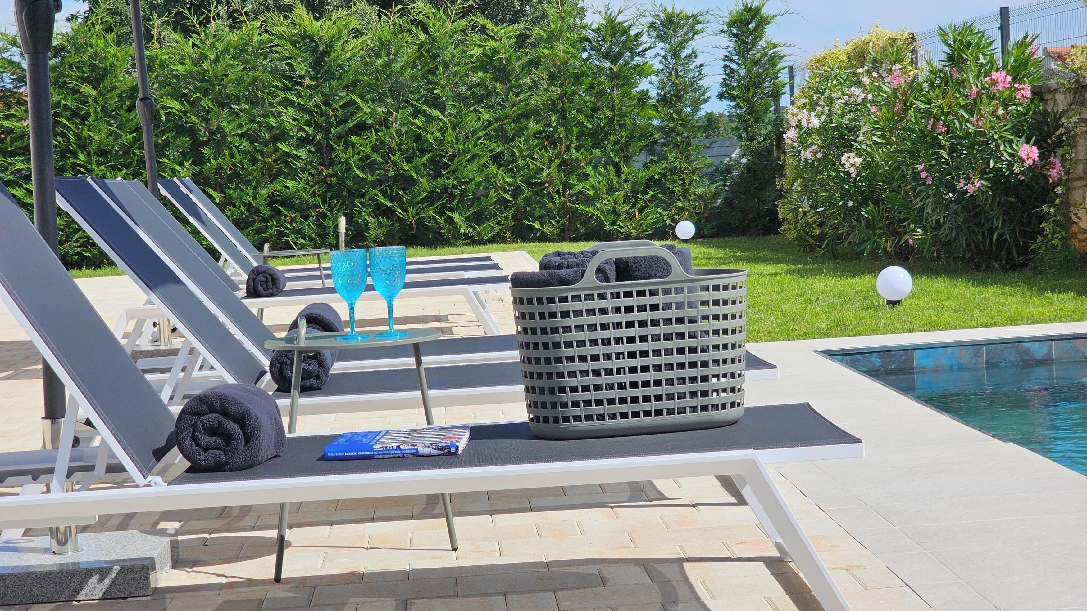 Un área junto a la piscina con sillones de descanso, una mesa con dos vasos azules, una cesta y un libro.