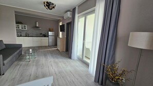 A modern living room with a grey sofa, a glass coffee table, and a lamp. There is a kitchen area with white cabinets and a grey countertop. A sliding glass door leads to a balcony with grey curtains. - Apartments with Shared Pool Casa Bianca (Kanfanar)