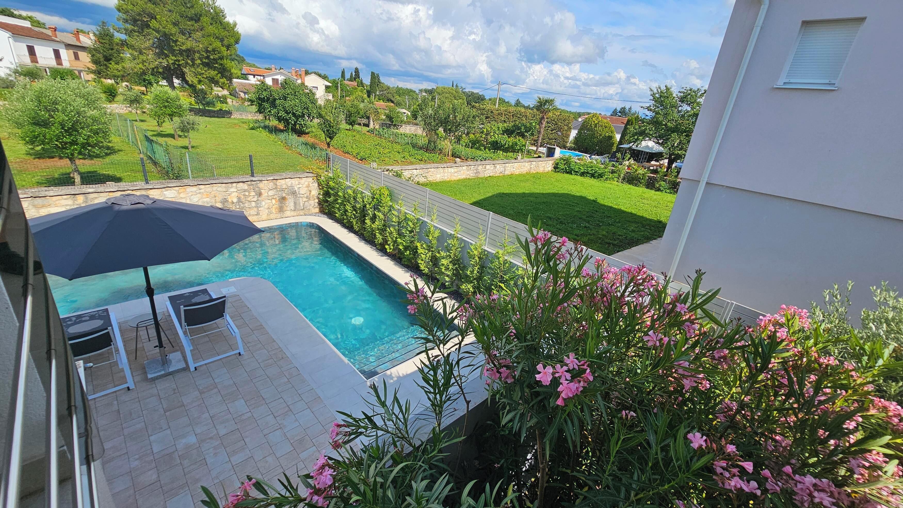 Une piscine avec deux chaises longues et un grand parasol, entourée d’un mur de pierre et d’une végétation luxuriante.