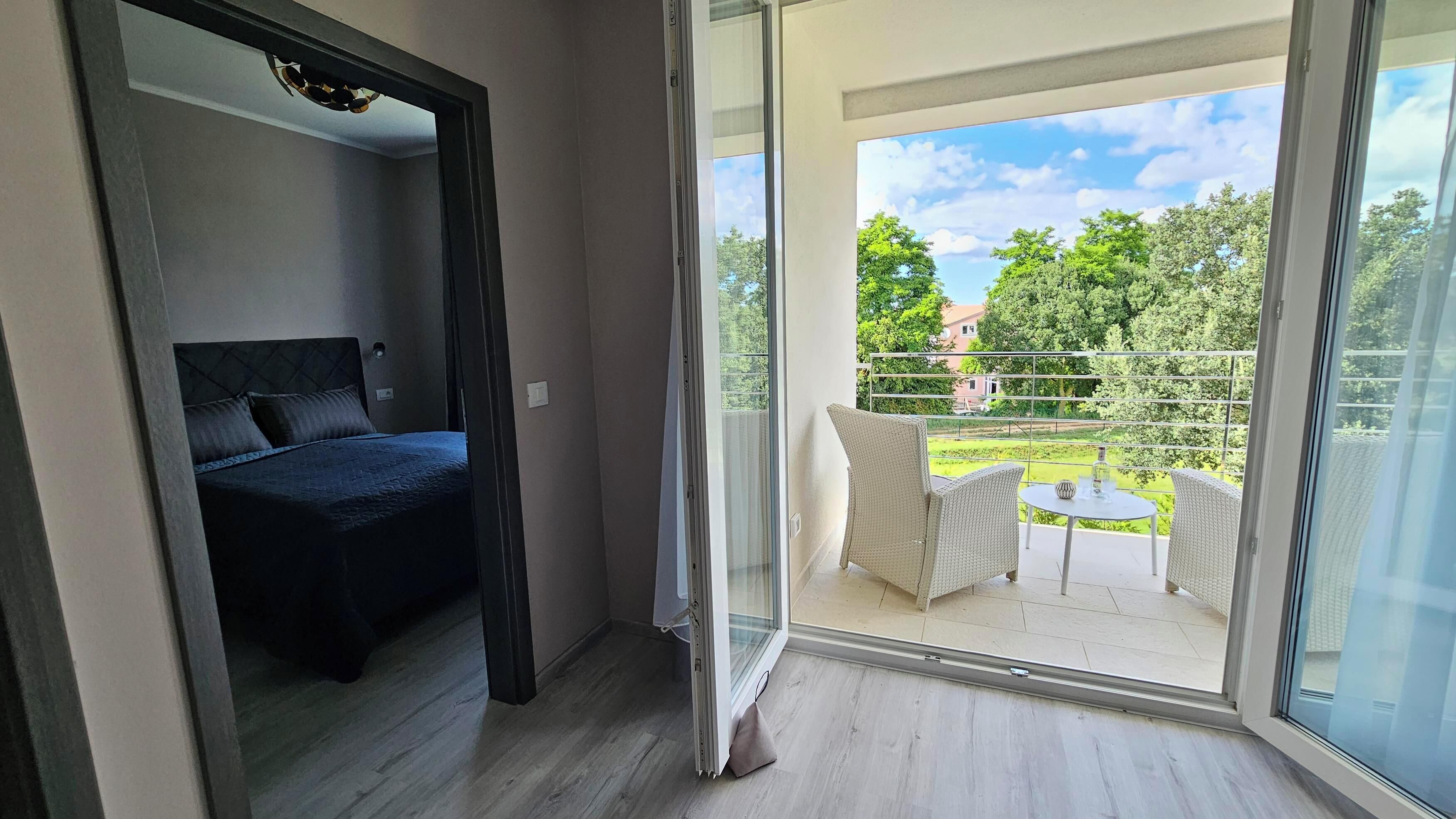 A bedroom with a bed and a balcony with a view of greenery and a house.
