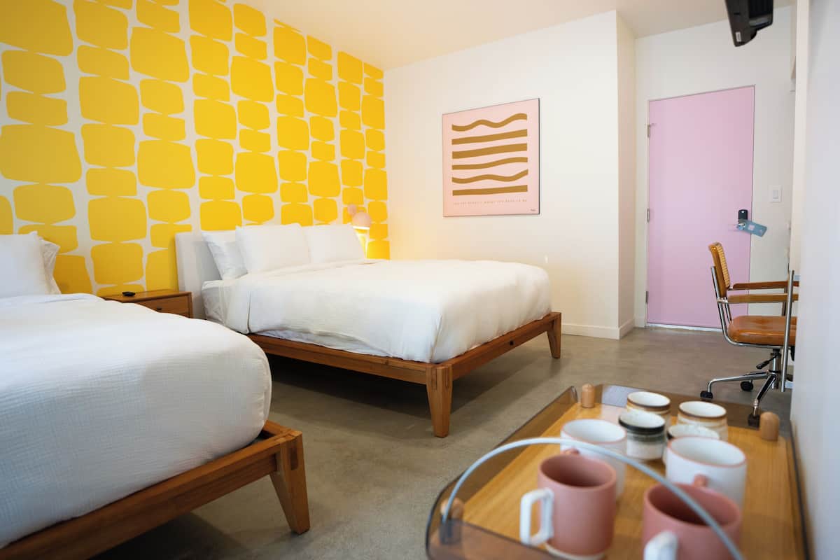 Habitación de hotel con dos camas, una mesita de noche de madera, un escritorio con una silla y una puerta rosa.
