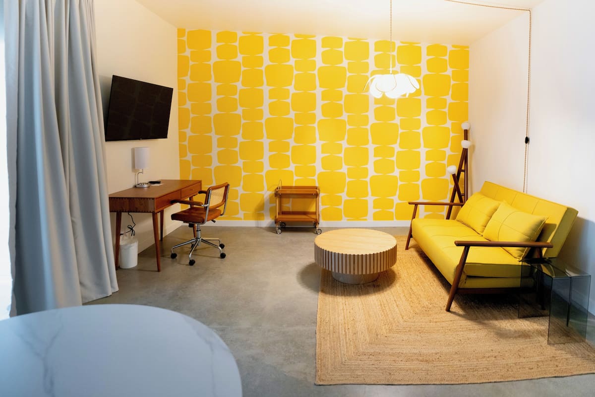 Una habitación con pared amarilla estampada, un escritorio de madera, un sofá amarillo, un otoman redondos y un televisor montado en la pared.