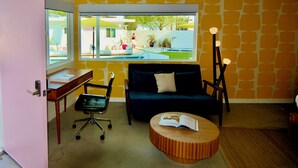 Une pièce comprenant un bureau, un fauteuil de bureau noir, un canapé bleu foncé, une table basse ronde en bois et une grande fenêtre donnant sur une piscine et un espace verdoyant.