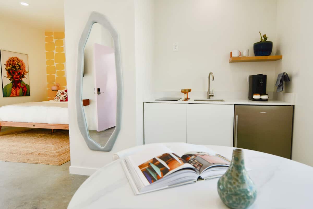 Una habitación de hotel moderna con una cama, una mesa redonda con un libro y un florero, un espejo, un lavabo, una cafetera y una estantería con una planta y tazas.