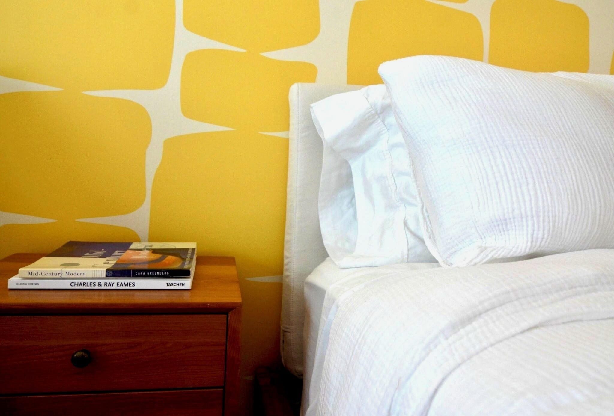 Una cama bien hecha con ropa blanca y almohadas, una mesita de noche de madera con algunos libros, y una pared amarilla con un diseño geométrico.
