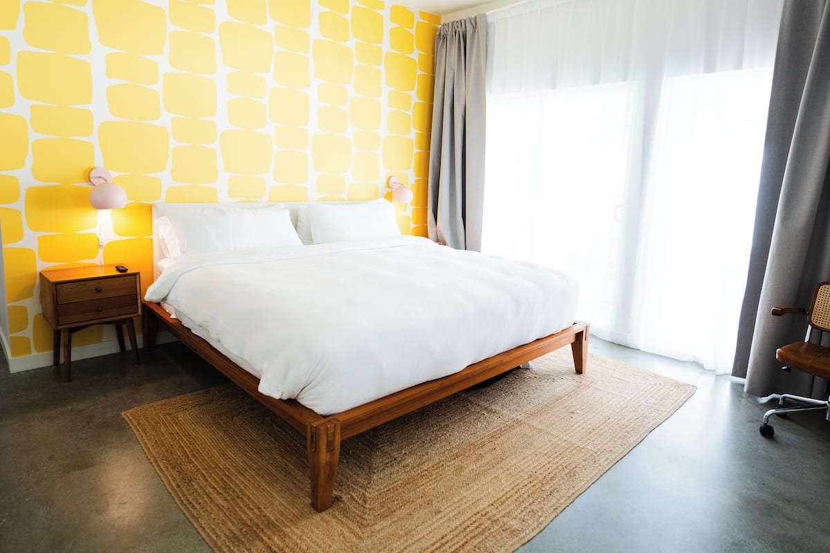 Un dormitorio con cama, mesita de noche, lámpara, silla y ventana con cortinas.