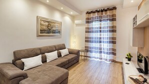 Ein Wohnzimmer mit braunem Sofa, einem gerahmten Bild an der Wand, einem gestreiften Vorhang und einem Flachbildfernseher auf einem Schrank.
