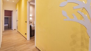 Ein Flur mit Holzboden, einer gelb strukturierten Wand und einer weißen Tür, die zu einem Zimmer mit Bett und Schreibtisch führt.