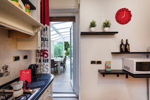 A modern kitchen with a gas stove, a microwave, and a red clock on the wall. There are two potted plants and a shelf with bottles.