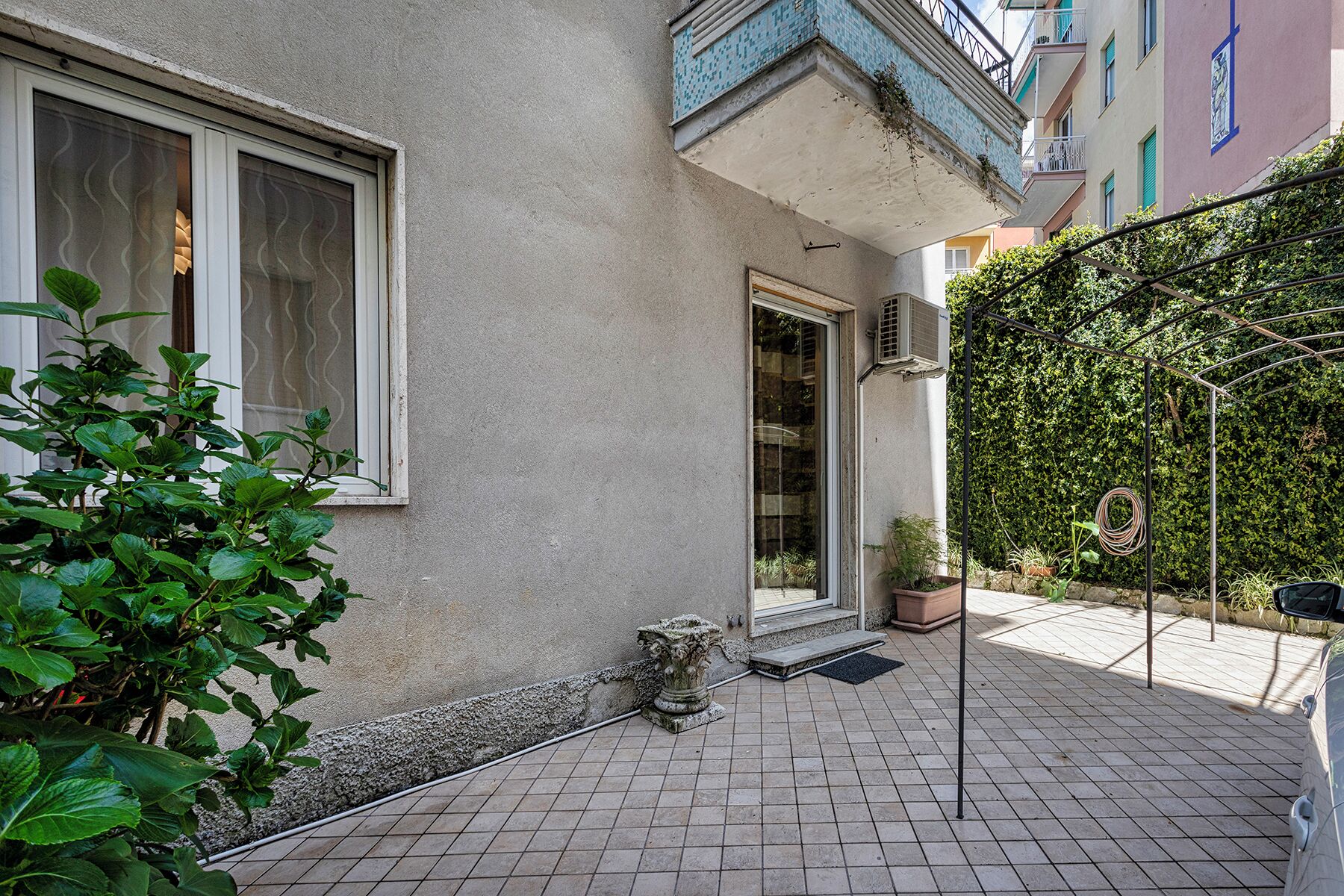 Un patio con suelo de baldosas, una planta en maceta, un jarro de piedra y una pérgola de metal.