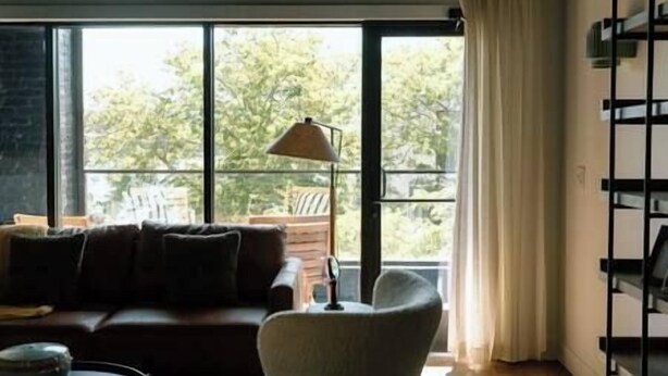 En moderne stue med sofa, lænestol og lampe. Store vinduer giver udsigt til træer.