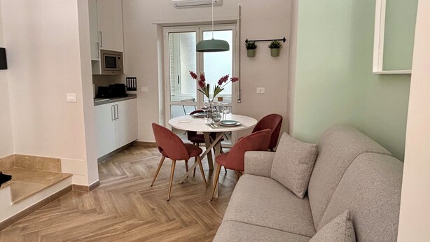 A modern living room with a beige sofa, a dining table with red chairs, and a kitchen area with white cabinets and a microwave.