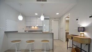 Une cuisine moderne avec trois tabourets de bar blancs, une table d’appoint en bois avec une lampe et des spots encastrés au plafond.