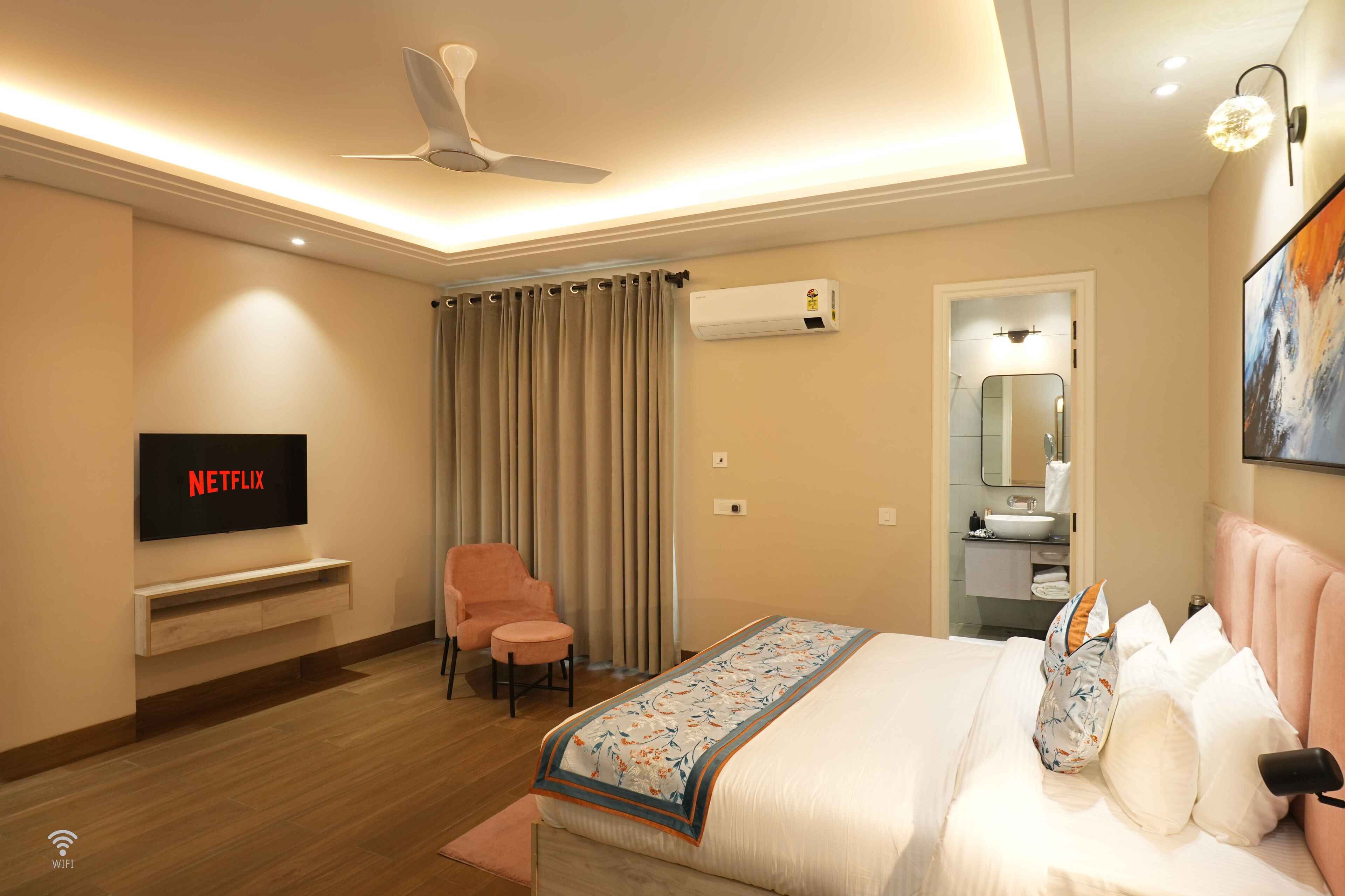 A modern hotel room with a bed, a TV displaying 'NETFLIX', a ceiling fan, an air conditioning unit, a bathroom with a mirror, and a painting on the wall.