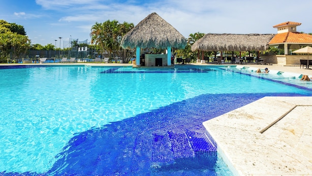 A swimming pool with a thatched-roof structure and a building with a red-tiled roof. - Family Elite Golf Vacations in Cocotal (Punta Cana)