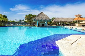 A swimming pool with a thatched-roof structure and a building with a red-tiled roof. - Family Elite Golf Vacations in Cocotal (Punta Cana)