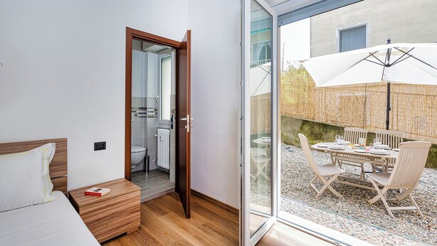 Une chambre à coucher avec un lit en bois, une table de chevet et une porte vitrée donnant sur un balcon équipé d’une table et de chaises.