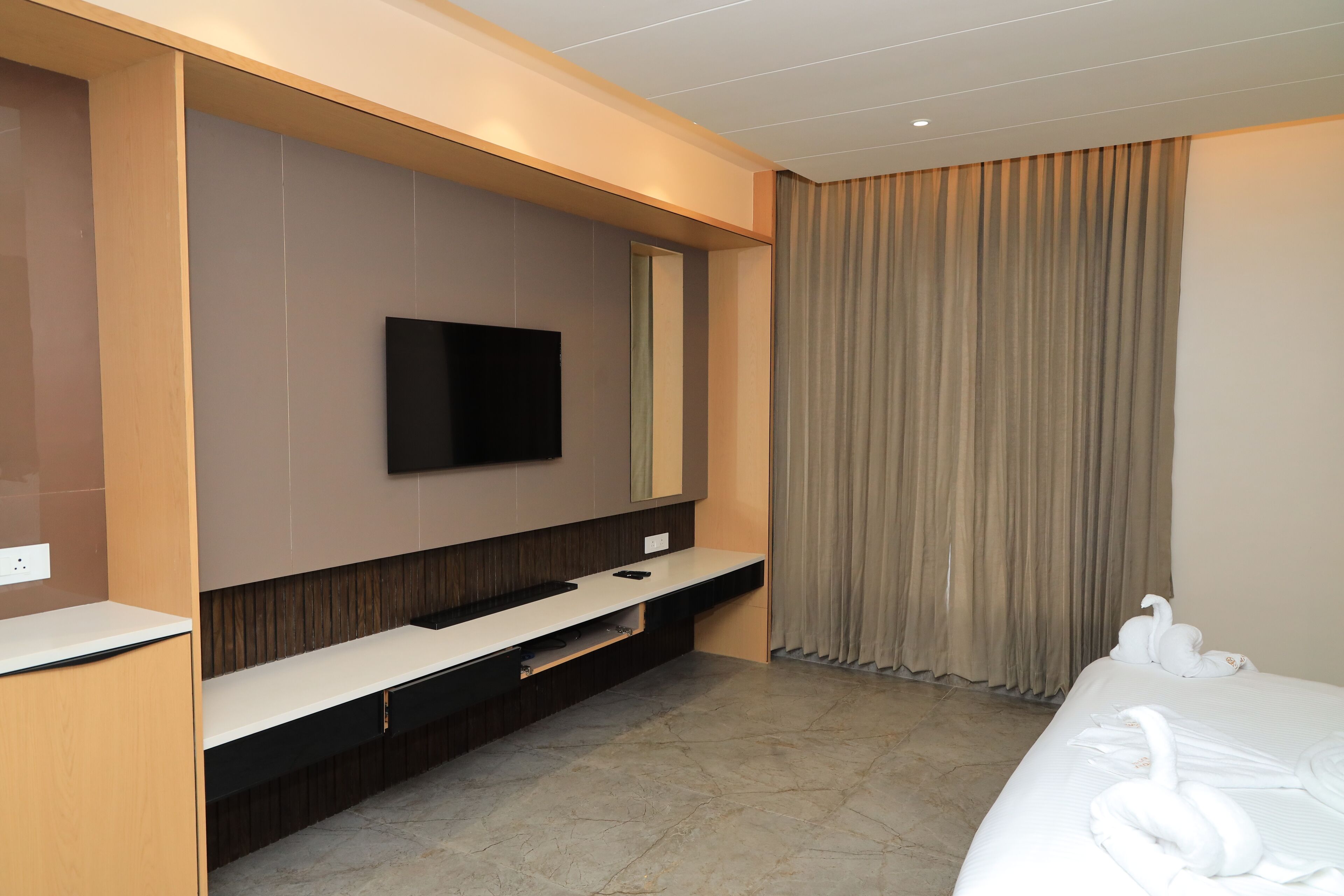 A modern hotel room with a flat-screen TV mounted on the wall, a built-in TV stand, a bed with white linens, and a large window with curtains.