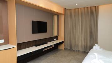 A modern hotel room with a flat-screen TV mounted on the wall, a built-in TV stand, a bed with white linens, and a large window with curtains.