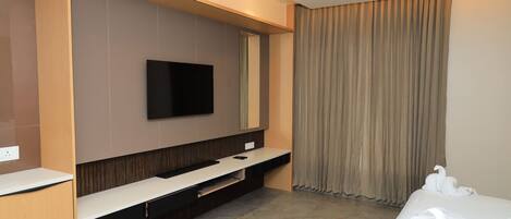 A modern hotel room with a flat-screen TV mounted on the wall, a built-in TV stand, a bed with white linens, and a large window with curtains.
