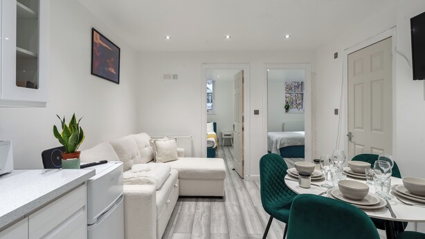 A modern, well-lit room with a kitchenette, a dining area with green chairs, and a cozy seating area with a sofa and cushions. - Beautiful Flat in London- Close to Central London- Canary Wharf- London ExCel (London)