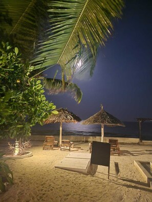 A beach with wooden lounge chairs and thatched umbrellas, a palm tree, and a view of the ocean at night.