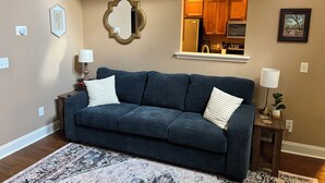 Ein Wohnzimmer mit einem dunkelblauen Sofa, einem gemusterten Teppich, einem Spiegel und einer durch einen Bogen sichtbaren Küche.