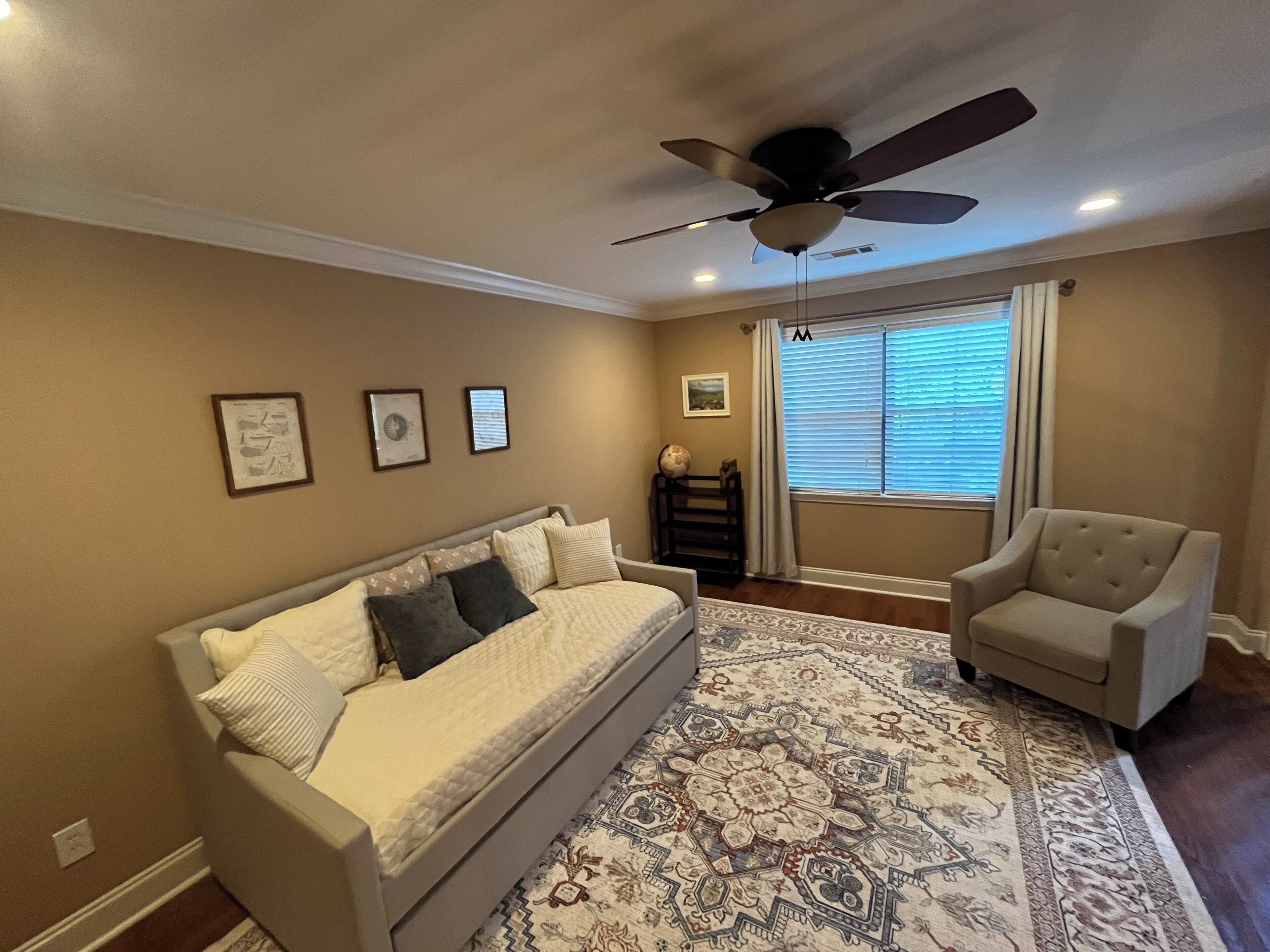 A living room with a beige sofa, a patterned rug, a ceiling fan, and a window with blinds.