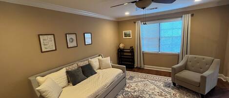 A living room with a beige sofa, a patterned rug, a ceiling fan, and a window with blinds.