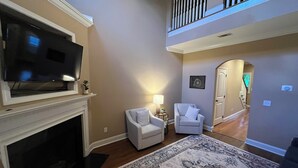 Ein Wohnzimmer mit Kamin, Fernseher, zwei weißen Sesseln, einem gemusterten Teppich und einer Treppe, die in einen oberen Bereich führt.