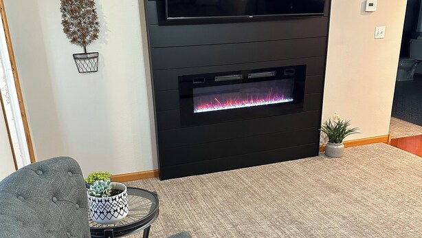 A living room with a flat-screen TV mounted above a fireplace, a grey sofa, and a small table with a potted plant. - Cozy 2-bd home in Newfolden. WiFi, AC, Full kitchen & living room w fireplace!  (Newfolden)