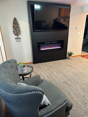 Un salon avec un téléviseur à écran plat fixé au-dessus d’une cheminée, un canapé gris et une petite table sur laquelle est posée une plante en pot.