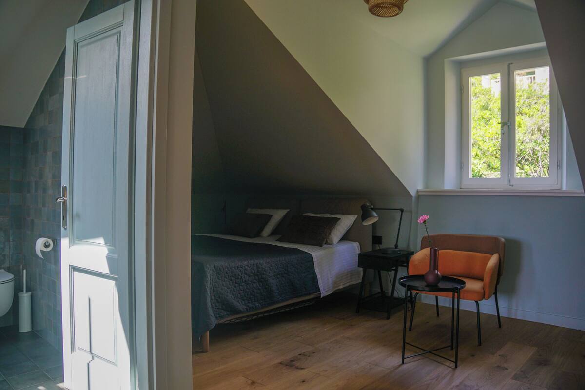 A bedroom with a sloped ceiling, a bed, a chair, and a window.