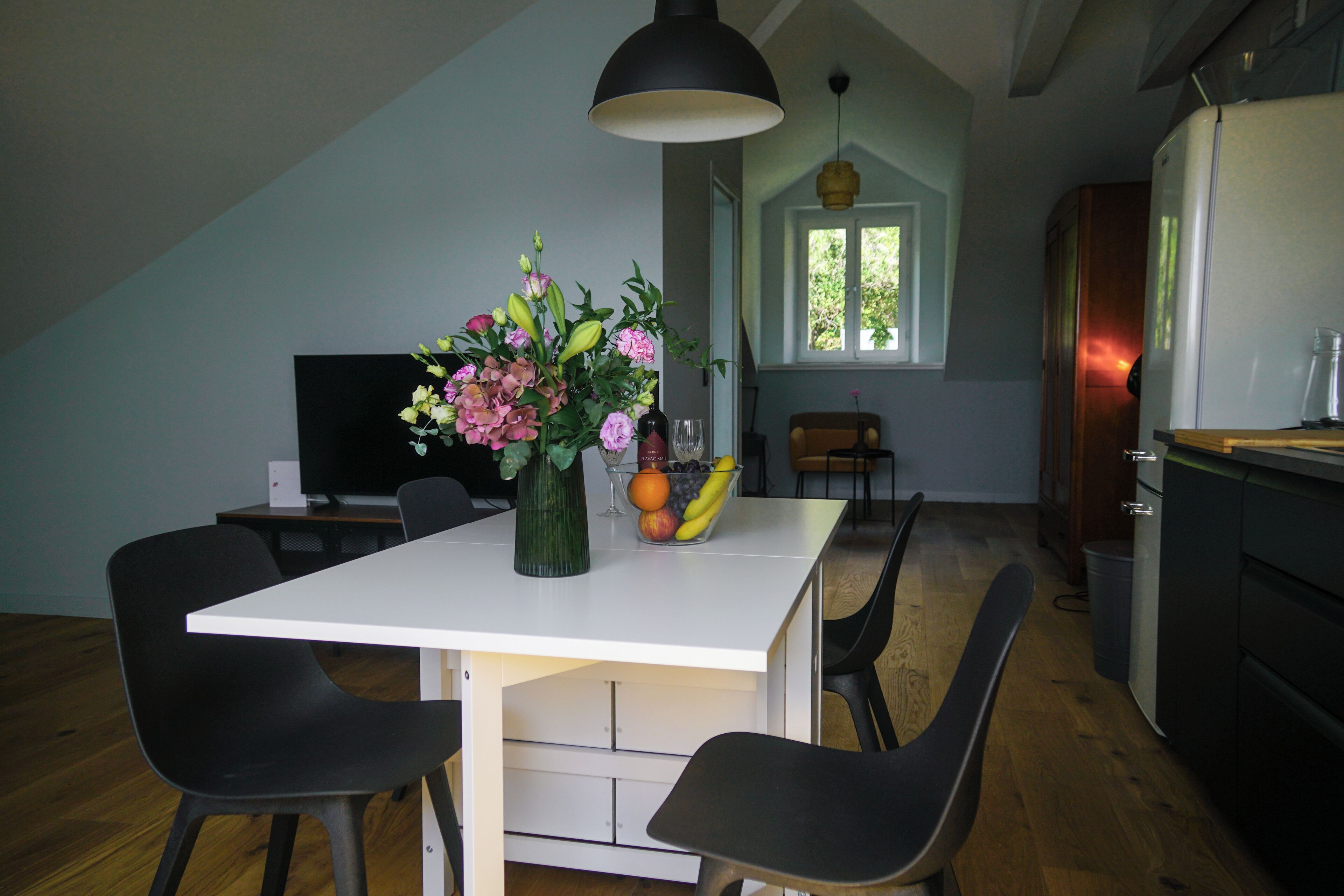 Ein Essbereich mit einem weißen Tisch, schwarzen Stühlen, einer Blumenvase und einer Obstschale.