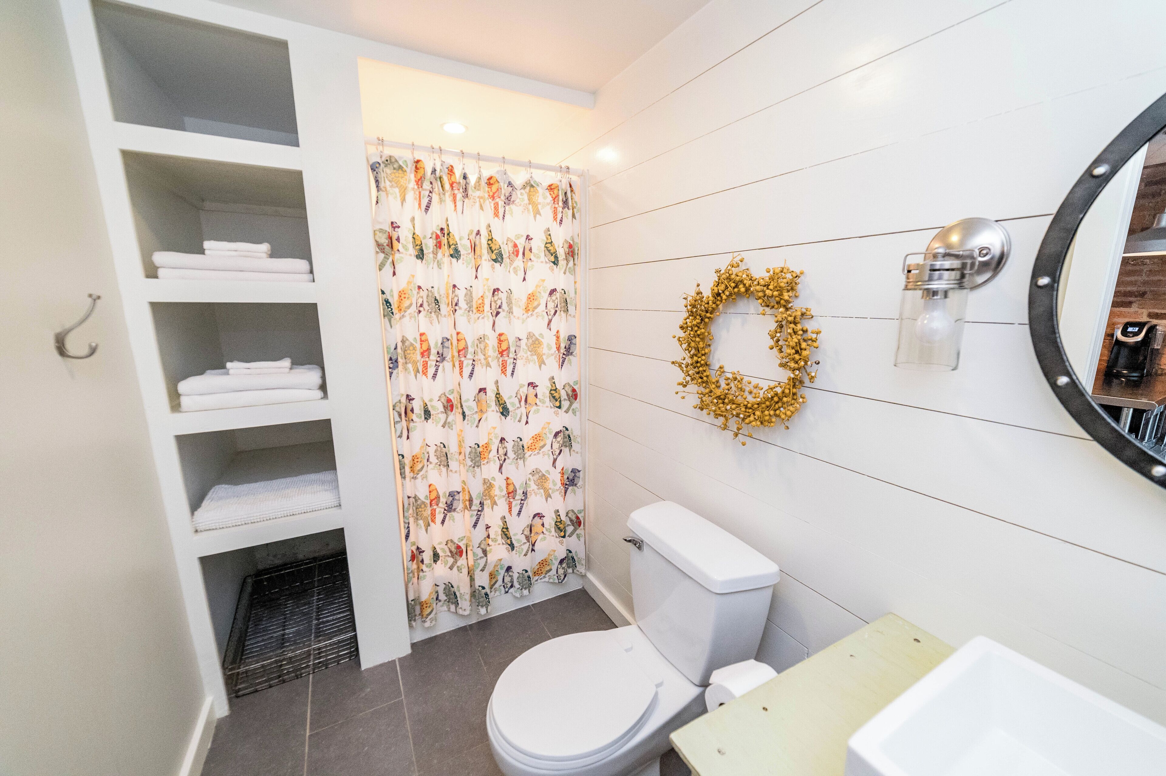 Ein Badezimmer mit Toilette, Waschbecken, einer Duschkabine mit Vogelmuster, einem weißen Regal und einer Wandleuchte.