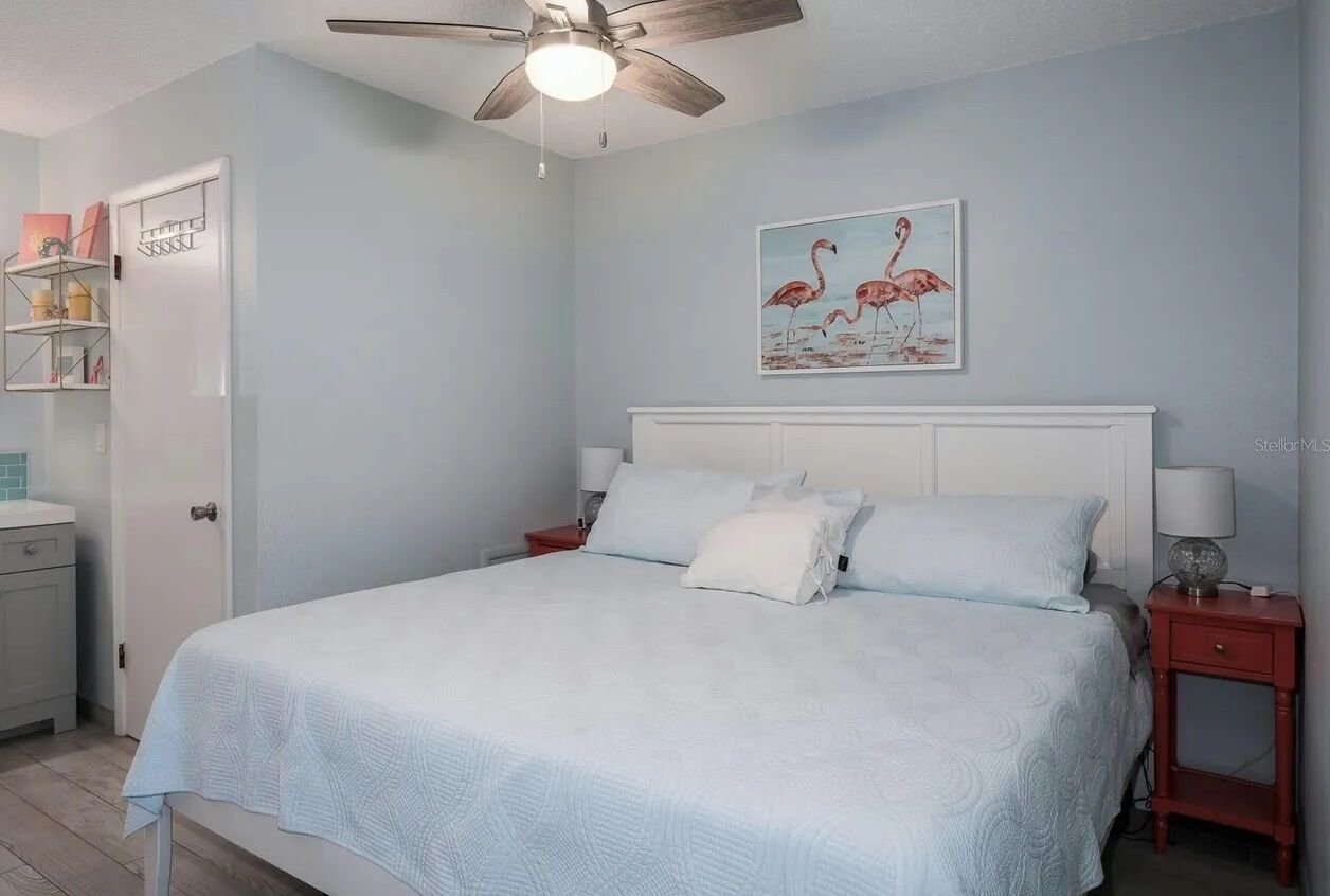 Une chambre à coucher comprenant un lit, un ventilateur de plafond, une table de chevet, une petite table d’appoint rouge et une étagère fixée au mur.