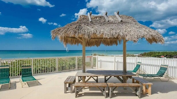 Eine Terrasse am Strand mit einem Reetdachschirm, hölzernen Bänken und grünen Liegestühlen.
