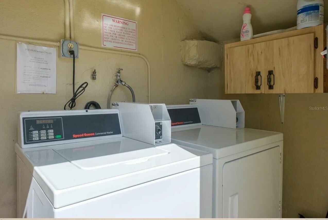 Un espace buanderie compact comprenant deux lave-linge professionnels de marque Speed Queen, un meuble contenant des produits de nettoyage et un panneau d’avertissement concernant l’utilisation de la lessive.
