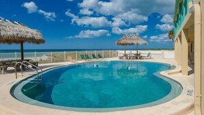 Ein klarer, blauer Pool mit geschwungener Kante, umgeben von Liegestühlen und Reetdachschirmen, mit Blick auf den Ozean und einem Gebäude im Hintergrund.